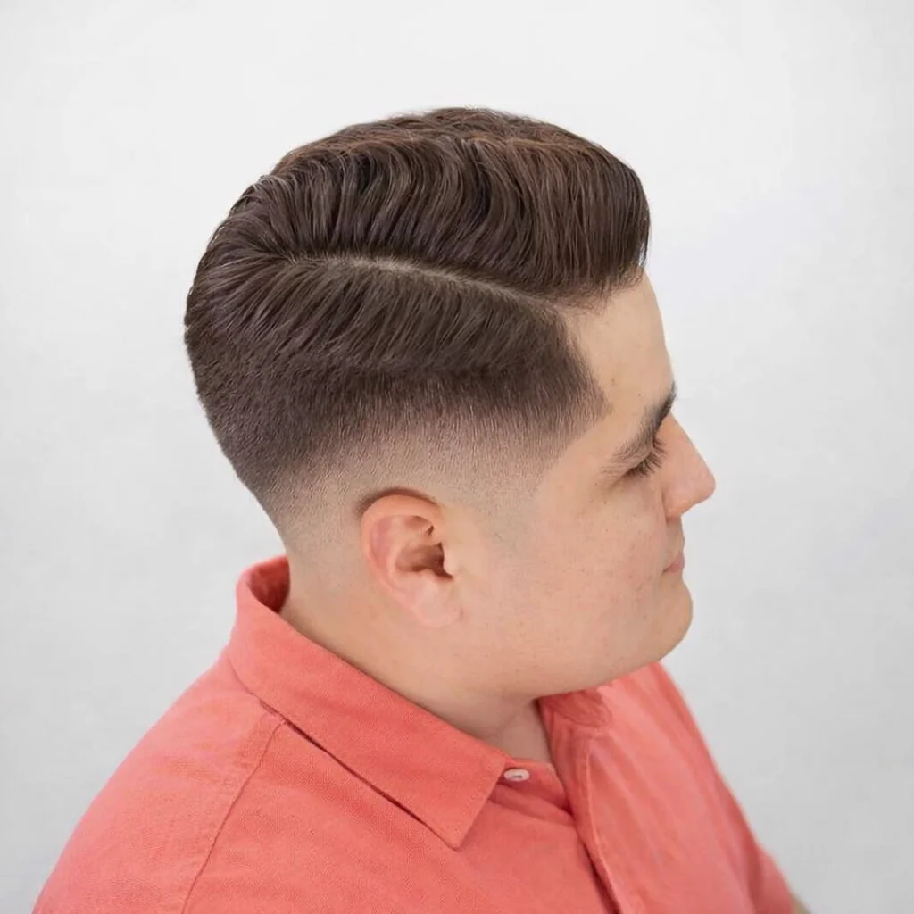 Potongan rambut mid fade yang halus dengan belahan samping dan gaya comb-over bertekstur, ditata rapi pada rambut pendek berwarna gelap dengan latar belakang putih bersih.

