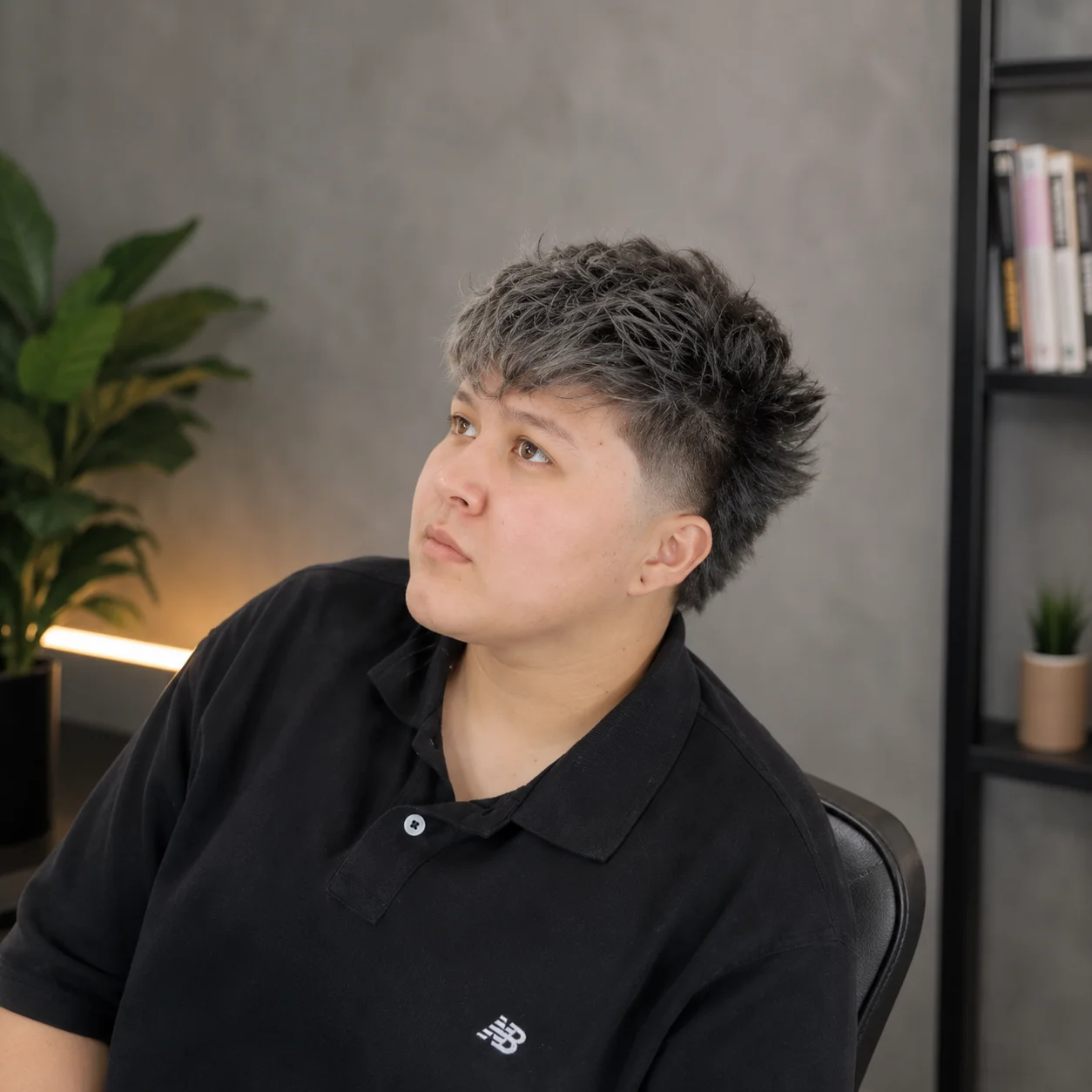 Potongan rambut pendek bertekstur dengan sisi ber-fade pada seorang individu muda yang memakai baju polo hitam, melihat sedikit ke atas dalam suasana studio dalaman moden dengan latar belakang kabur yang lembut.