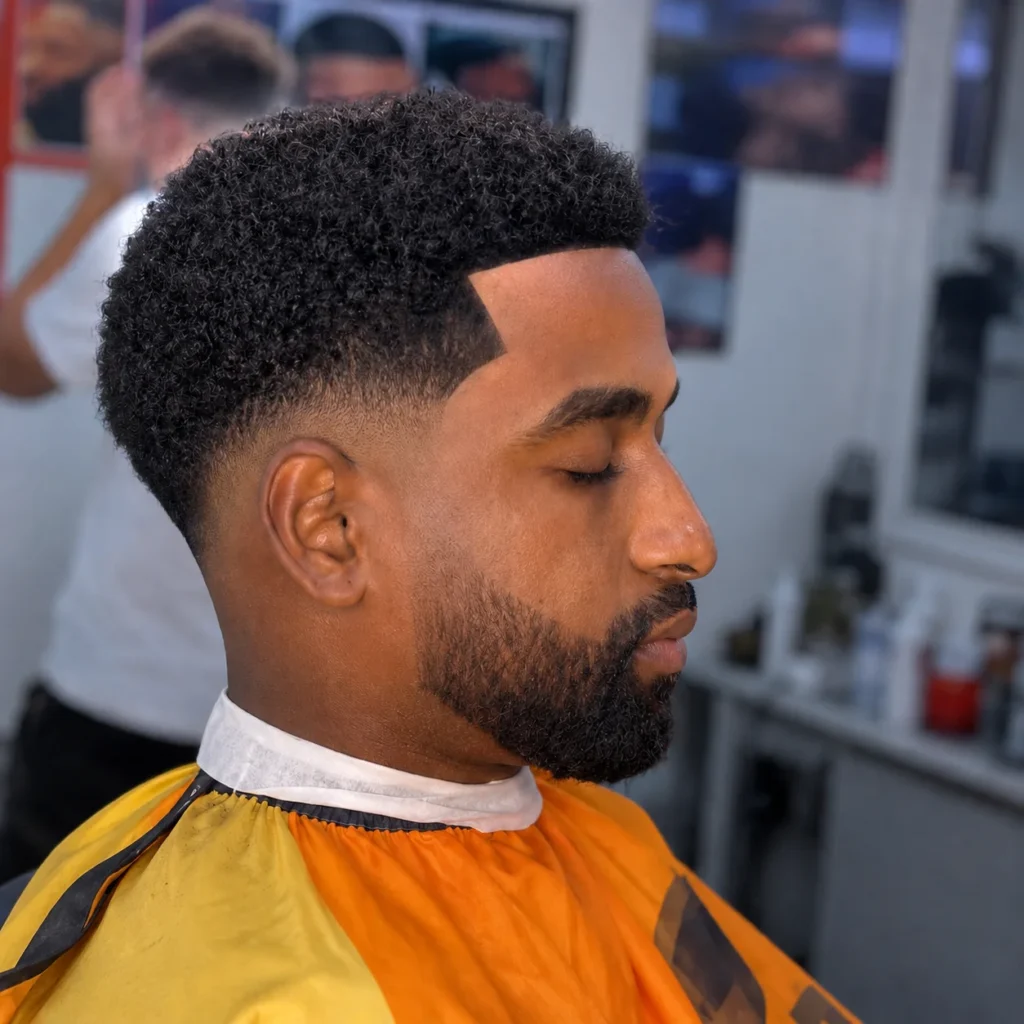 Potongan rambut low fade tajam dengan bagian atas rambut keriting bertekstur dan garis janggut yang rapi, diambil di suasana barbershop modern.
