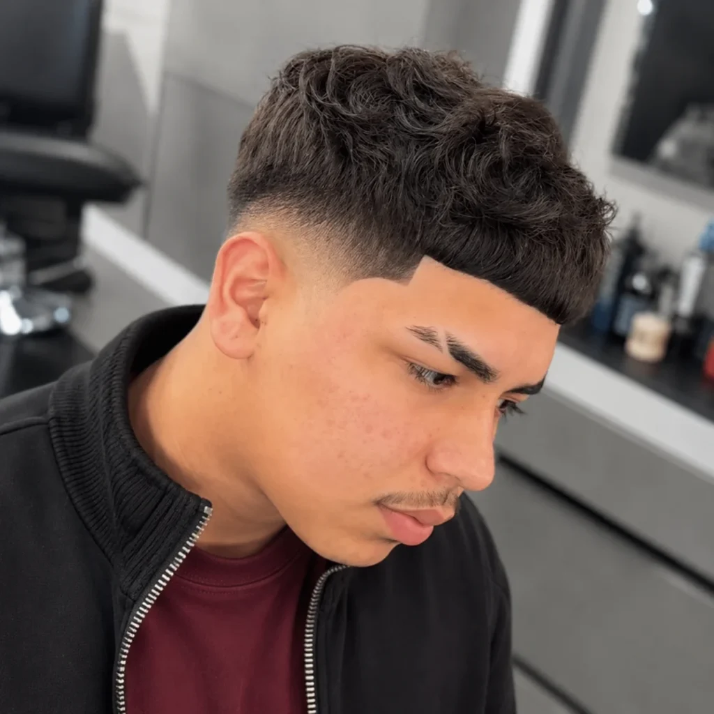 Pria muda dengan potongan rambut curly Edgar fade, mengenakan jaket hitam di barbershop modern.
