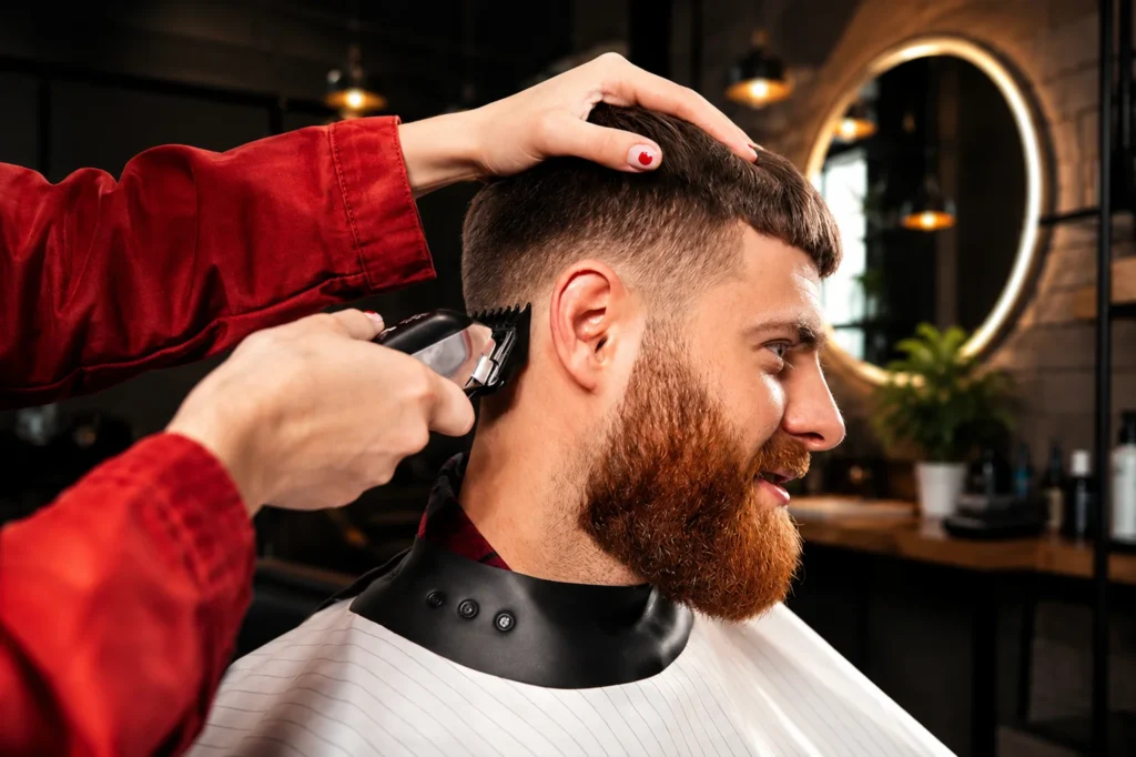 Professional barber using clippers to create a clean fade haircut on a bearded man in a modern luxury barbershop with warm lighting.